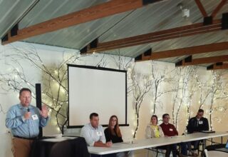 Five people are seated at a table with a projector screen behind them, and one man is standing at the podium speaking with a microphone.
