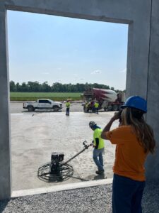A young woman in a hard hat looks out a window at a construction site.