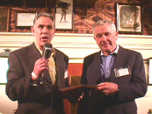 Two men in suits, one holding a microphone and the other holding an award plaque.