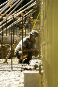 A construction worker crouched, measuring a concrete wall in a construction site.