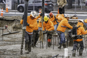 Nine construction workers, eight of which in flourescent orange hoodies and hard hats, pound rebar into the ground in a construction site in the rain.