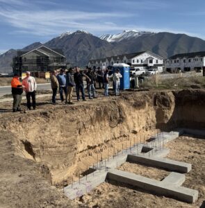 A group of people stand on the ground looking at the beginning of a concrete foundation below ground-level.