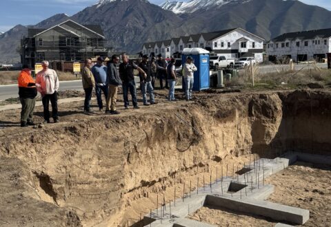 A group of people stand on the ground looking at the beginning of a concrete foundation below ground-level.