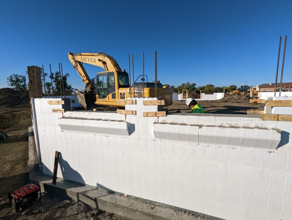 Side view of white concrete foundation walls with an excavator above and a construction worker behind the wall.