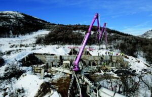 A pink crane extends over a concrete foundation mid-construction. Built in the hills with snow and trees around. People stand atop the wall.