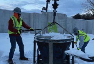 Two construction workers are working on a piece of machinery in the snow.
