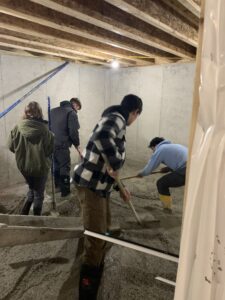 Four students are spreading gravel in a concrete room that is under construction.