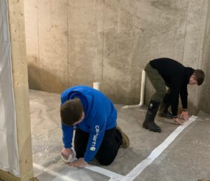 Two students are taping down plastic sheeting in a concrete room under construction.
