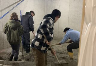 Four students are spreading gravel in a concrete room that is under construction.