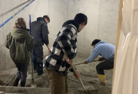 Four students are spreading gravel in a concrete room that is under construction.