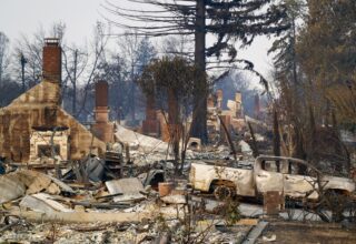 A house surrounded by trees that has been destroyed by a fire. There is burnt debris all around the ground.