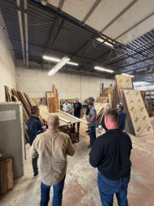 Nine men stand around a table with two white boards on it in a warehouse of sheeting material. One person films on his phone.