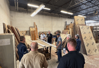 Nine men stand around a table with two white boards on it in a warehouse of sheeting material. One person films on his phone.